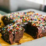 A close-up of a vegan cosmic brownie with a bite taken out to reveal the dense, fudgy interior, topped with a chocolate ganache and colorful sprinkles.
