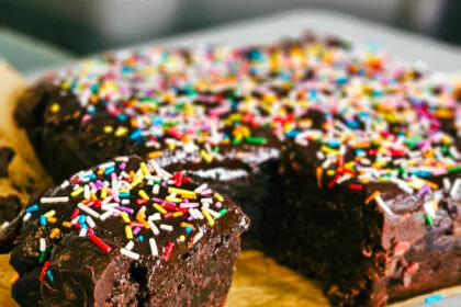 A close-up of a vegan cosmic brownie with a bite taken out to reveal the dense, fudgy interior, topped with a chocolate ganache and colorful sprinkles.