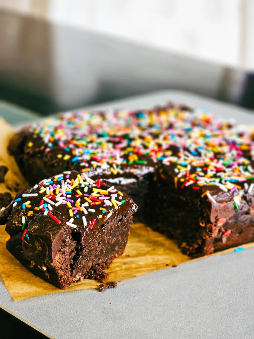 A close-up of a vegan cosmic brownie with a bite taken out to reveal the dense, fudgy interior, topped with a chocolate ganache and colorful sprinkles.