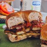 A delectable Charred Tofu Bacon Sandwich cut in half, showcasing layers of golden-brown tofu, vegan bacon, melted vegan cheese, and fresh veggies inside a toasted whole grain bagel.