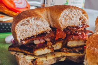 A delectable Charred Tofu Bacon Sandwich cut in half, showcasing layers of golden-brown tofu, vegan bacon, melted vegan cheese, and fresh veggies inside a toasted whole grain bagel.