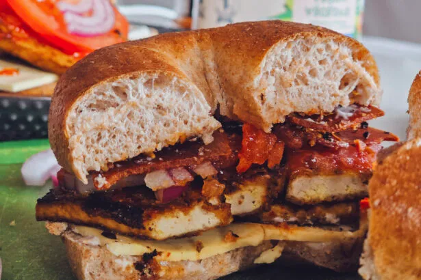 A delectable Charred Tofu Bacon Sandwich cut in half, showcasing layers of golden-brown tofu, vegan bacon, melted vegan cheese, and fresh veggies inside a toasted whole grain bagel.
