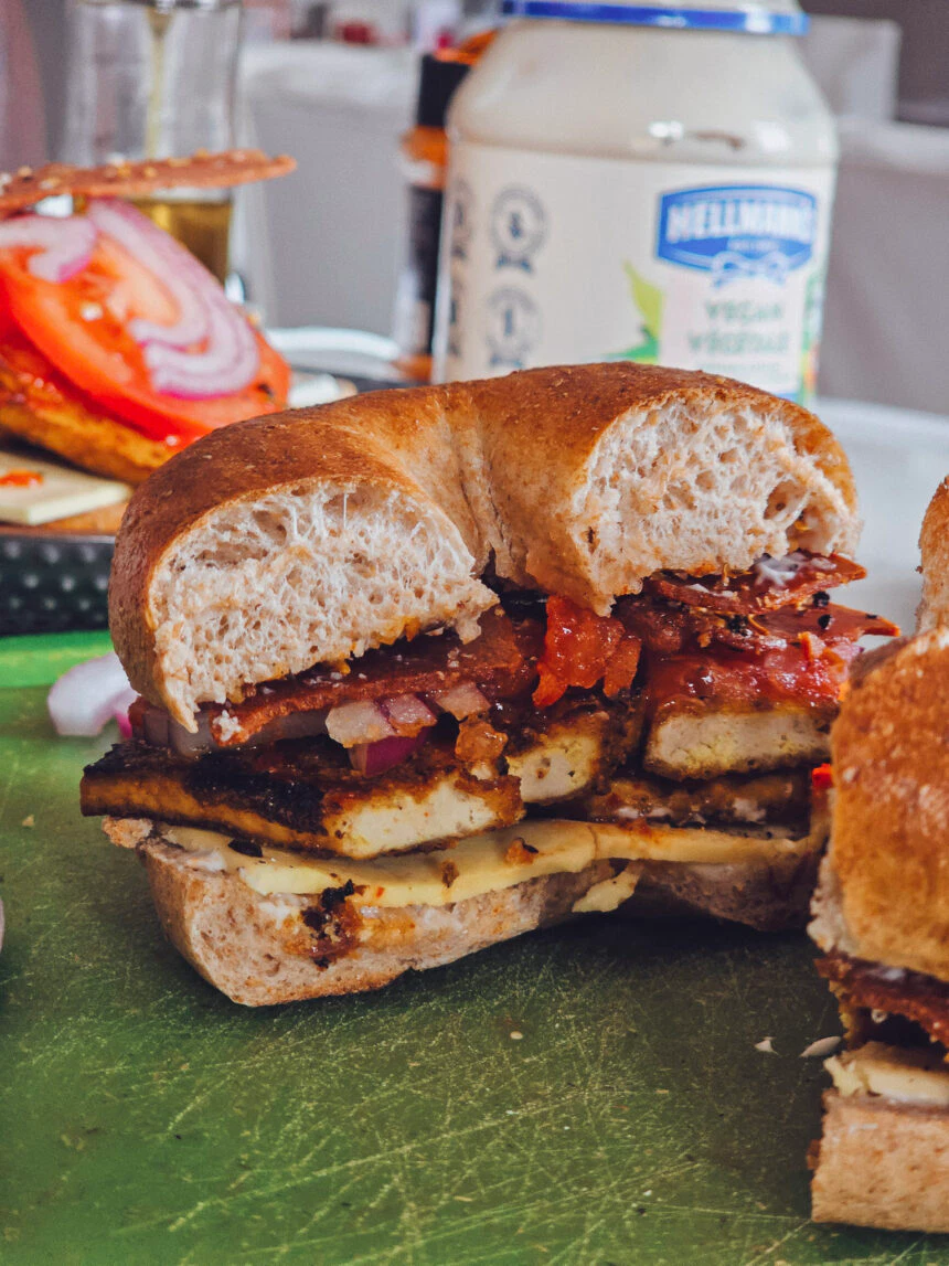 A delectable Charred Tofu Bacon Sandwich cut in half, showcasing layers of golden-brown tofu, vegan bacon, melted vegan cheese, and fresh veggies inside a toasted whole grain bagel.